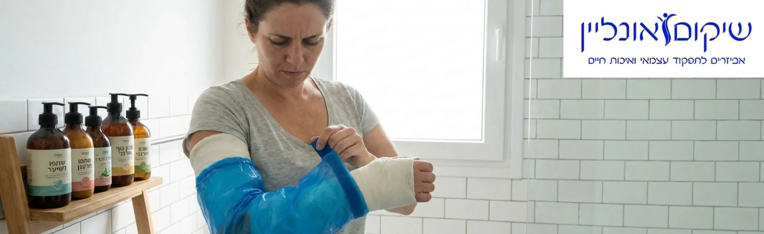 אישה עם גבס ביד ימין עומדת בחדר אמבטיה מואר, כשהיא מכסה את הגבס שלה בשרוול פלסטיק אטום למים כהכנה למקלחת. על המדף לצידה מונחים בקבוקי שמפו עם משאבה.